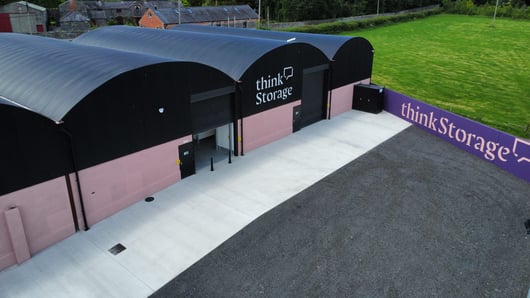 Aerial view of a modern storage building with black curved roofs and light pink walls. The words 'think Storage' are displayed on the building and a nearby wall. Surrounding area includes a green grassy field and some older buildings in the background.