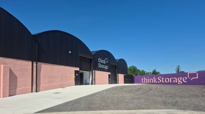 Wide angle shot of gravel drive way facing towards a barn-like building with a black arched roof. The front of the building is a light pink with large black roller shutter doors. There a is a large sign on the front of the building that says ‘think Storage’.